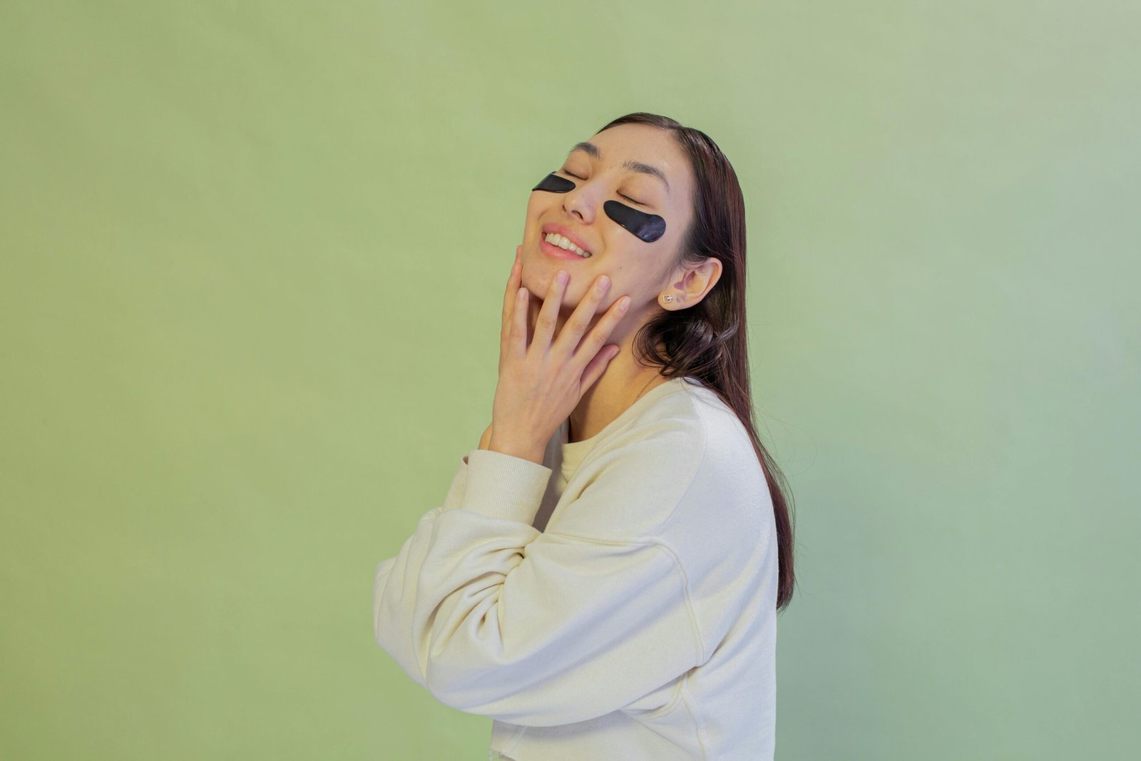 Young woman enjoying skincare routine with black hydrogel eye patches indoors, smiling and relaxed.